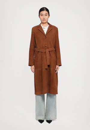 Donna che indossa un cappotto in suede marrone con cintura, jeans a gamba larga blu chiaro, scarpe nere a punta, in piedi di fronte a uno sfondo bianco semplice.