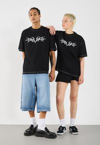 Black oversized t-shirts with white graphic design, paired with light blue denim shorts and black footwear. Model poses against a white background.