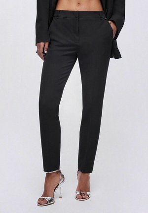 Donna che indossa pantaloni neri su misura a vita alta con dettaglio tasche, abbinati a sandali argentati con tacco alto e punta aperta, in piedi con le mani nelle tasche.