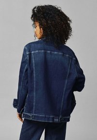 Giacca in denim scuro, con una vestibilità rilassata, cuciture in evidenza, dettagli con bottoni sulle maniche e un'etichetta all'orlo inferiore.