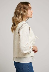 Femme aux cheveux blonds ondulés portant un blouse à manches longues couleur crème avec des volants bleus et un gilet matelassé, associé à un jean bleu.