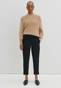 Pull en beige moelleux à manches longues associé à un pantalon noir ajusté et des mules noires, sur un fond minimaliste clair.
