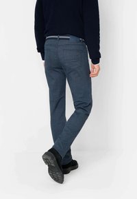 Blaue gewobene Hose mit slim fit, ausgestattet mit zwei Gesäßtaschen und einer strukturierten Oberfläche, kombiniert mit schwarzem Schuhwerk und einem navyblauen Oberteil.