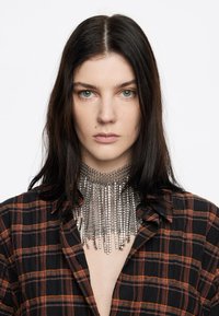 Collana choker in argento con design a frange sovrapposte, abbinata a una camicia a quadri nera e arancione. Focus sulla texture metallica e sul motivo geometrico.