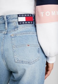 Jeans de denim azul claro con un parche azul oscuro visible etiquetado como "TOMMY JEANS" y una pequeña bandera de logo en el bolsillo trasero.