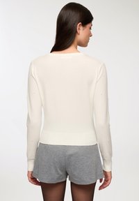 Maglione bianco lavorato a maglia con orlo a costine e maniche lunghe, decorato con piccoli dettagli sulle spalle. Abbinato a pantaloni corti grigi.