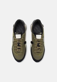 Baskets vert olive avec une tige texturée, des accents noirs, des coutures jaunes et une semelle intérieure à motif. Comprend des lacets et une languette de tirage.