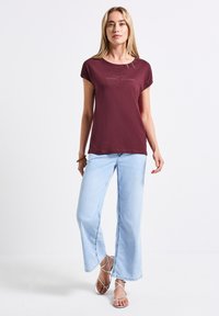 Camiseta corta de manga en burdeos con cuello redondo y texto, combinada con jeans de pierna ancha de color azul claro y sandalias plateadas con tiras.