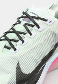 Chaussure de course verte et noire avec un dessus texturé, des accents blancs, de grosses lacets et un détail rose sur la semelle. Présente un logo Nike bien en vue.