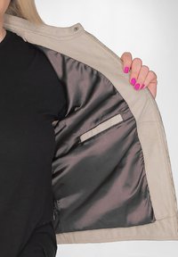 Beige jacka med en glänsande mörk foder, med en sidficka i fodret. Tyget verkar slätt och något texturerat.