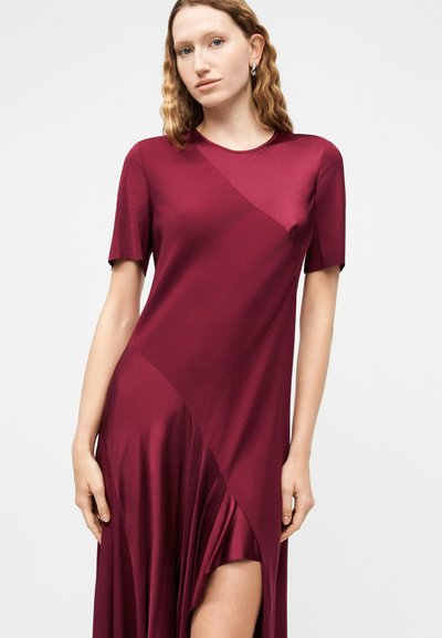 Γυναίκα με κυματιστά ξανθά μαλλιά που φοράει ασημένια σκουλαρίκια και ένα ασύμμετρο, μπουρντό σατέν φόρεμα με κοντά μανίκια και ψηλό σκίσιμο στο μηρό.