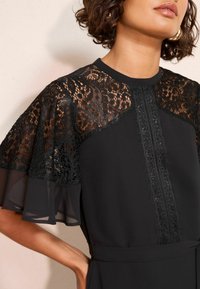 Čierne šaty s priesvitnými čipkovými rukávmi a detailmi. Majú okrúhly výstrih a textúrovaný pásik na prednej strane. Hladký povrch látky.