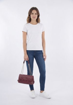 Femme portant un t-shirt blanc, un jean bleu, des baskets blanches, tenant un sac à main matelassé bordeaux avec des anses en chaîne sur un fond uni.
