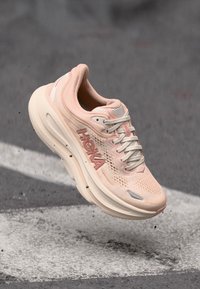 HOKA tekaških čevljev z zgornjim delom iz svetlo rožnatega mrežastega materiala, teksturiranimi sloji, belim srednjim delom in ravnimi vezalkami. Zračno blaženje je vidno na podplatu.