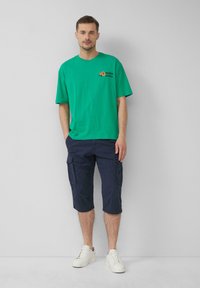T-shirt en coton vert avec un motif orange, associé à un short cargo navy avec des poches, et des baskets blanches. Fond uni.