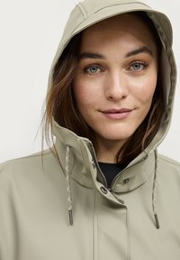 Beige waterdichte jas met een capuchon, ritssluiting en trekkoorden. Beschikt over een zachte textuur en zilveren accenten op de trekkoorden.