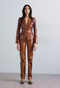 Costum din piele maro, cu o jachetă ajustată și pantaloni care prezintă un model subtil în formă de semilună. Textură netedă, detaliu cu un singur buton și pantofi verzi.
