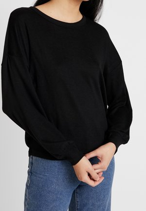 Frau trägt ein locker sitzendes schwarzes Langarmshirt mit rundem Ausschnitt und blaue Jeans, die Hände locker vor dem Körper verschränkt.