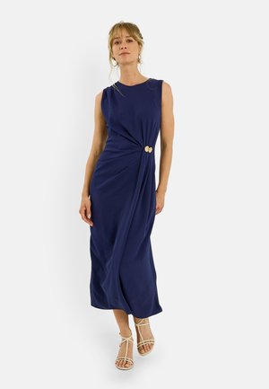 Mujer que lleva un vestido midi sin mangas azul marino con un nudo lateral y sandalias beige con tiras sobre un fondo liso.