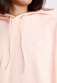 Ljusrosa hoodie med snörning och logotyp. Tyget är mjukt och något texturerat. Har en enkel design med en vit Nike-emblem på bröstet.