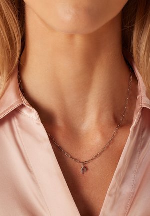 Zilveren ketting met een langwerpig linkontwerp, voorzien van een kleine 'P' hanger, gedragen tegen een lichtroze satijnen shirt.