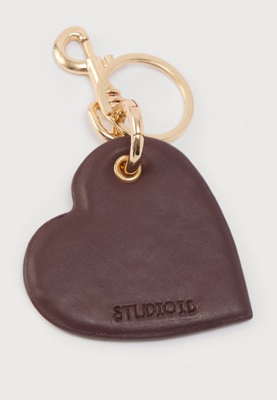 Καφέ δερμάτινο μπρελόκ σε σχήμα καρδιάς με χρυσό μεταλλικό κούμπωμα και κρίκο, με χαραγμένο το κείμενο "STUDIOD" στην επιφάνεια.