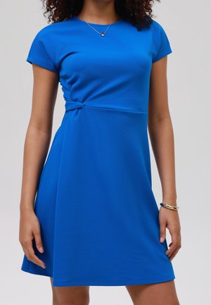 Kobieta ubrana w krótką sukienkę o długości do kolan w jasnoniebieskim kolorze z detalem węzła z boku, naszyjnikiem i bransoletką na jednolitym tle.