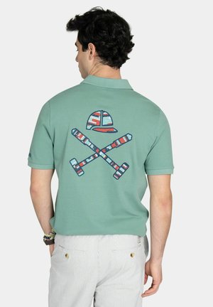 Hombre vestido con una camiseta tipo polo verde con un diseño en la espalda de mazos cruzados en rojo, blanco y azul, junto con una gorra, combinado con pantalones de rayas claras.