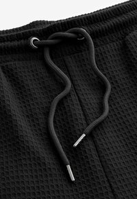 Tissu noir à texture gaufrée avec une taille à cordon de serrage. Le cordon est épais, arrondi et doté d'aglets en métal, améliorant la fonctionnalité et l'ajustement.
