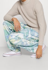 Person som bär en beige stickad tröja, blågrön byxor med vit virvelmönster, och vita sneakers, hukar med ena handen vilande på knäet.