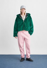Junge Frau trägt eine grüne, flauschige Zip-Hoodie, ein beiges Poloshirt, rosa Jogginghose, eine graue Mütze und schwarze Sneaker. Sie steht auf einem blauen Teppich.
