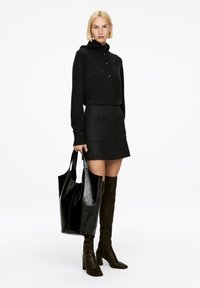 Schwarzer Strick-Cardigan mit Knöpfen, kombiniert mit einem dunkelgrauen Rock mit Fronttaschen. Das Model hält eine glänzende schwarze Tote-Bag und trägt braune kniehohe Stiefel.