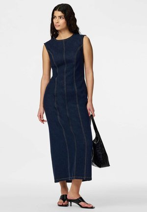 Vrouw met lang donker haar, die een mouwloze, aansluitende donkere denim midijurk draagt, zwarte sandalen met hakken, en een zwarte gestructureerde handtas vasthoudt.