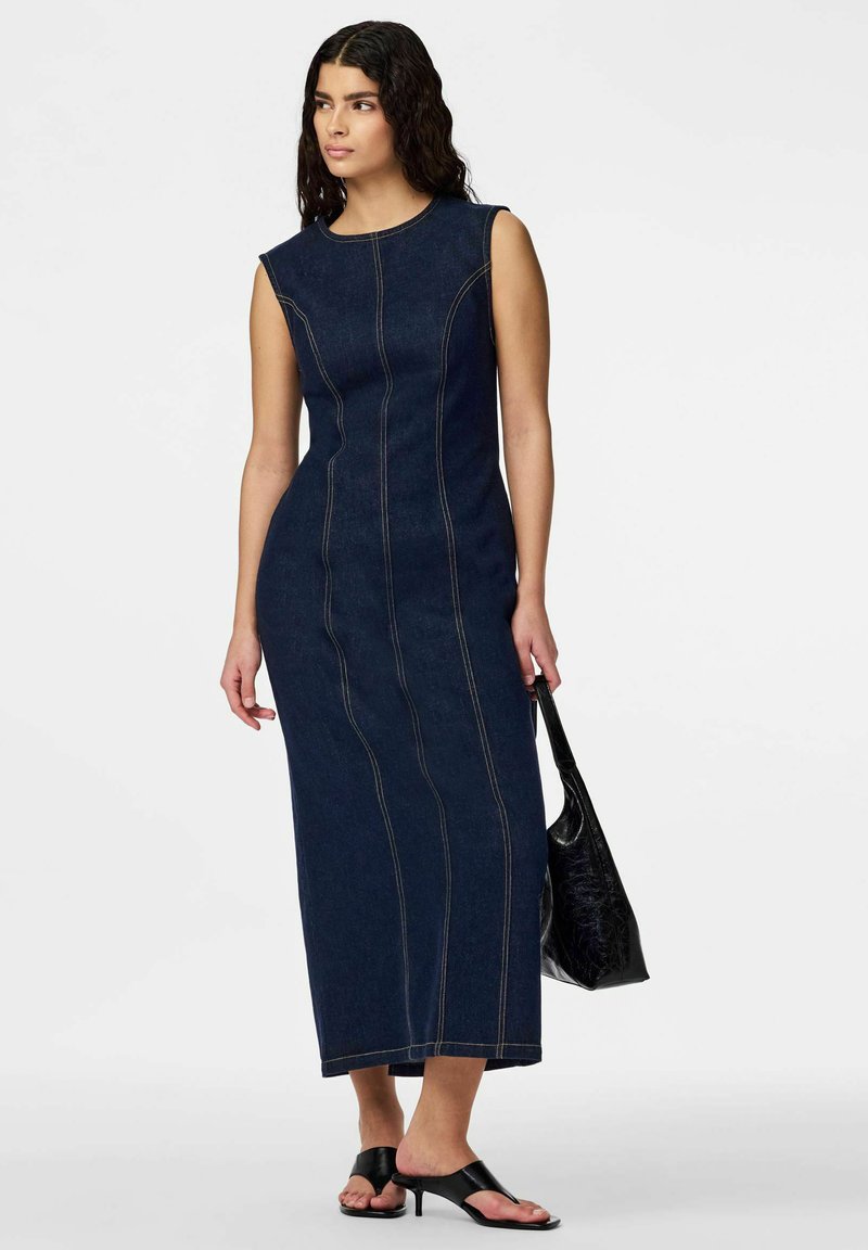 Γυναίκα με μακριά σκούρα μαλλιά που φοράει ένα αμάνικο εφαρμοστό σκούρο τζιν midi φόρεμα, μαύρα σανδάλια με τακούνι και κρατάει μια μαύρη υφασμάτινη τσάντα.