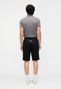 Pilka polo marškinėliai ir juodos šortai, pagaminti iš minkštos medžiagos. Šortai turi liemens etiketę; batai yra šviesiai kreminės spalvos. Vaizdas iš nugaros.