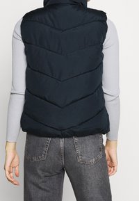 Granatowa pikowana kamizelka z wysokim kołnierzem, ozdobiona szwem w jodełkę i pikowaną fakturą, noszona na jasnoszarej bluzce z długim rękawem.