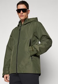 Olivgrüne, wasserdichte Jacke mit einem Vorderreißverschluss, verstellbarem Kapuze und Klettverschlussärmeln. Sie verfügt über zwei seitliche Taschen und ein schlankes Design.