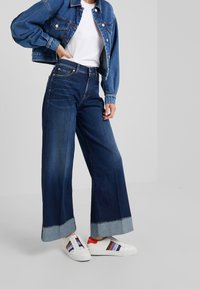 Kvinna som bär vida mörkblå jeans med uppvikta fållar, vit T-shirt, blå denimjacka och vita sneakers med röd häl och färgade ränder.