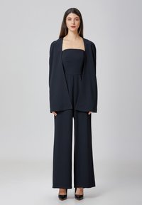Combinaison noire sans bretelles, avec un pantalon évasé et une veste ouverte et ample. Tissu lisse avec un minimum de détails.