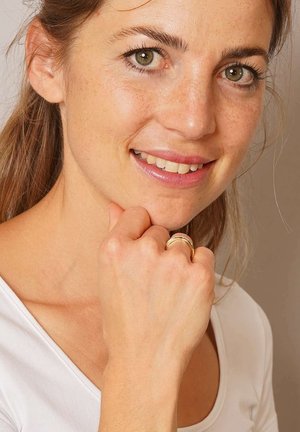 Goldring mit geschichtetem Design, glatter Textur und polierter Oberfläche an einer Hand, neben einem hellfarbenen Hemd auf einem neutralen Hintergrund.