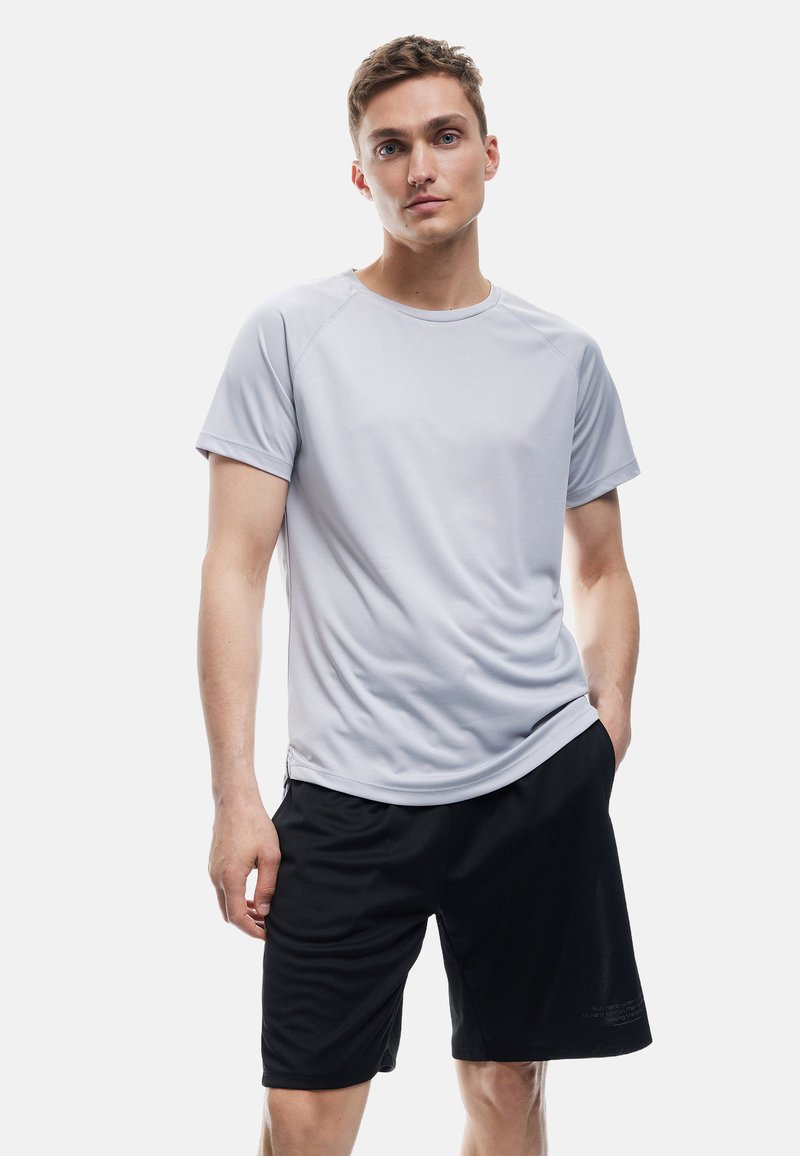 Junger Mann steht mit einer Hand in der Tasche, trägt ein hellgraues kurzärmliges Hemd und schwarze Sportshorts vor einem weißen Hintergrund.