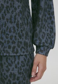 Il tessuto leopardato blu e nero presenta una texture morbida, maniche lunghe e polsini elasticizzati, con una mano visibile che afferra il capo al polso.
