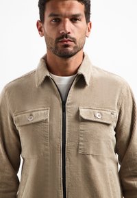 Man in een beige corduroy jacket met knoopzakken op de borst en een zwarte ritssluiting aan de voorkant over een wit shirt, kijkend naar voren.