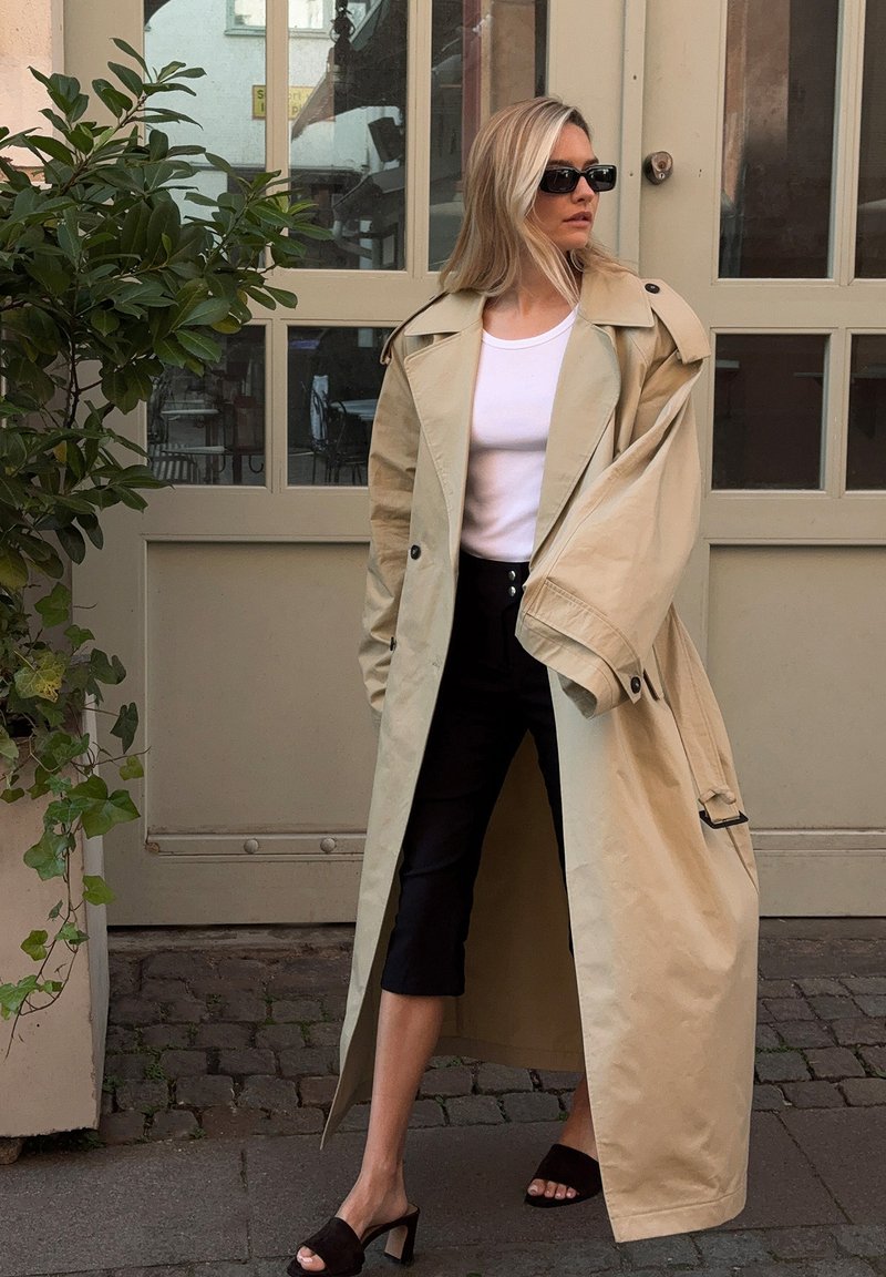 Femme blonde portant des lunettes de soleil noires, un haut blanc, un pantalon noir coupé, des sandales à talons noires et un trench beige surdimensionné, se tenant près des portes vitrées.