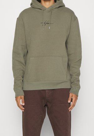 Person trägt olivgrünen Hoodie mit Fronttasche und schwarzem kursivem Text auf der Brust, kombiniert mit braunen Hosen.