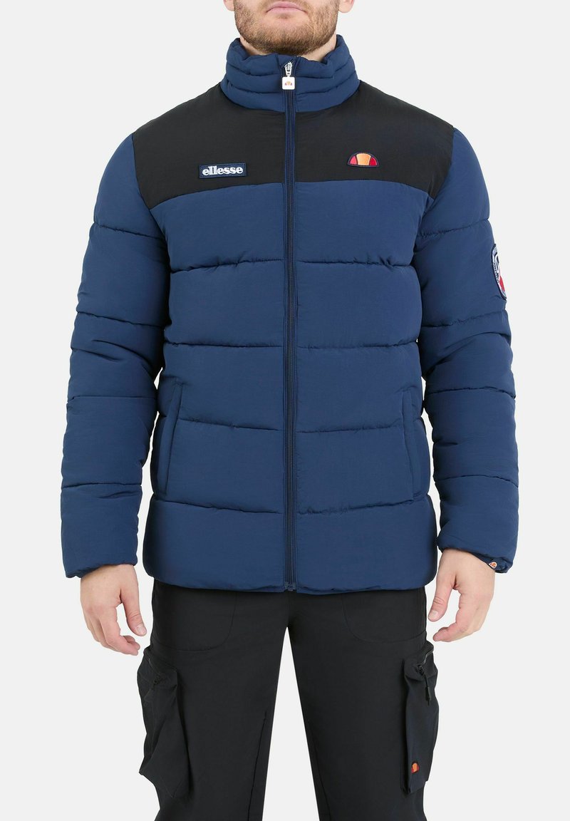 Giacca puffer blu navy con parte superiore nera, chiusura con zip e polsini elasticizzati. Presenta patch con logo sul petto e sulla manica sinistra.