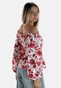 Witte off-shoulder blouse met rode bloemenpatronen, puffermouwen, geplooide details en een losse pasvorm, gecombineerd met een blauwe spijkerbroek.