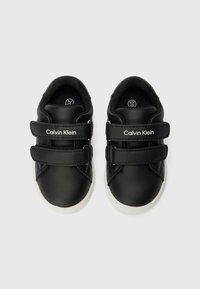 Sapatos de couro preto com duas fitas de Velcro, sola branca e logotipo em relevo "Calvin Klein" na lateral. Textura suave, forma arredondada.
