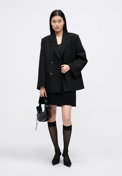 Blazer oversized preto de duplo peito, saia mini a condizer, meias até ao joelho pretas transparentes e sapatos de salto preto com bico pontiagudo. A segurar uma mala preta texturizada.
