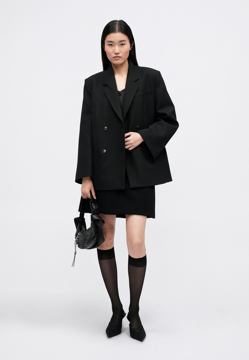 Black double-breasted oversized blazer, matching mini skirt, sheer black knee-high socks, and pointed-toe black heels. Carrying a textured black bag.